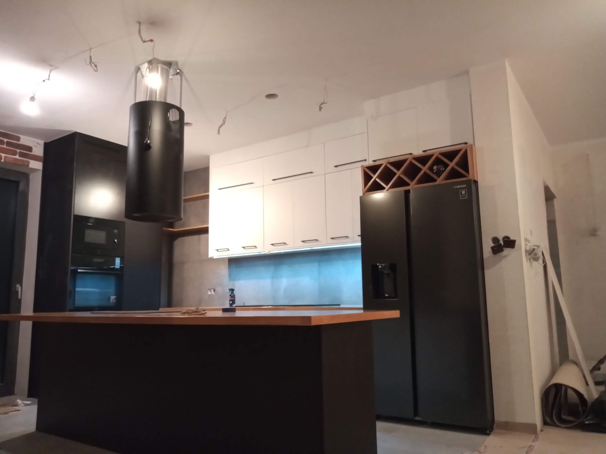 Nowoczesna kuchnia w trakcie montażu: czarne szafki, białe fronty, wyspa z drewnianym blatem, czarna lodówka i regał na wino. Widoczne niedokończone wykończenia.