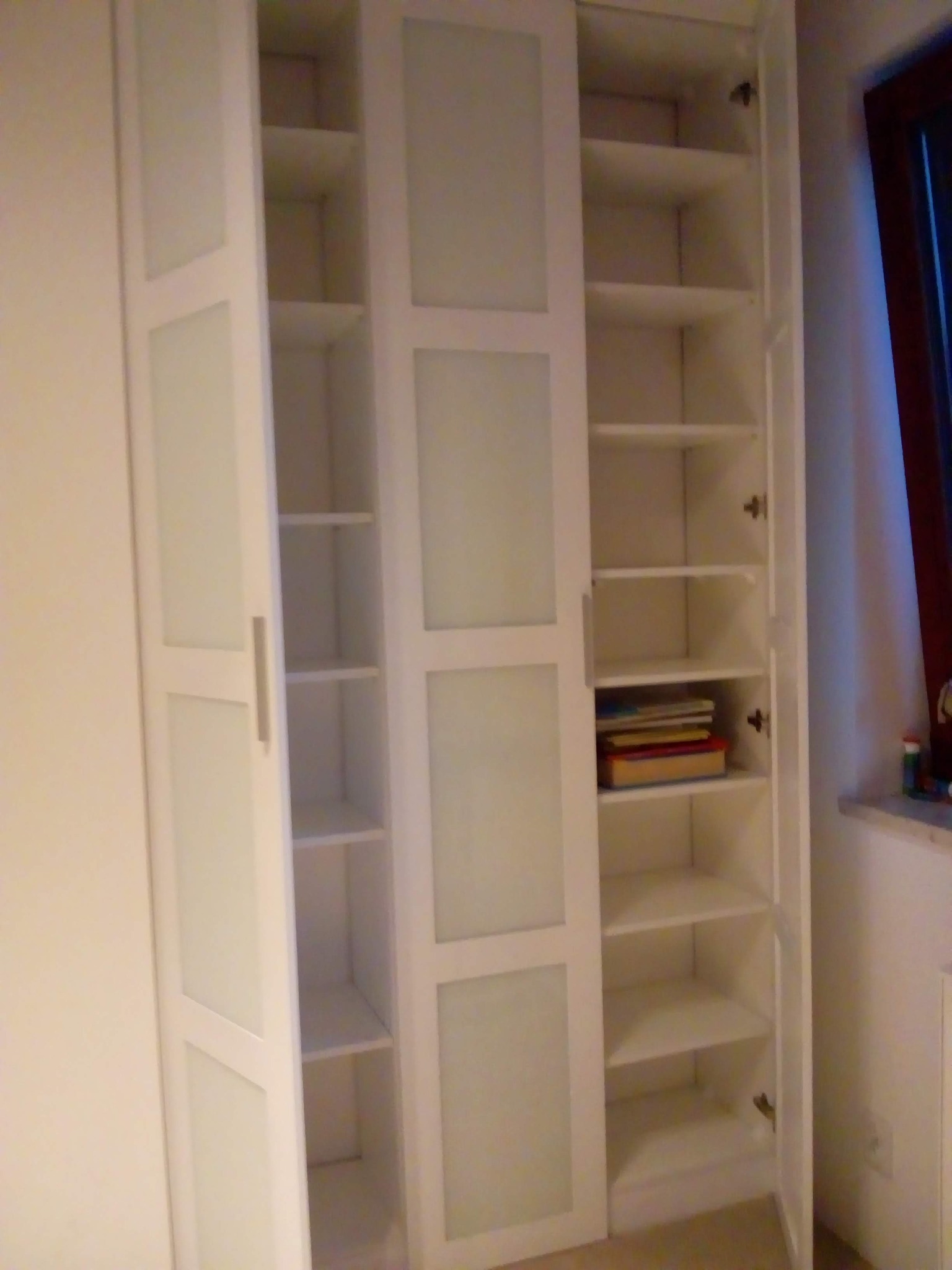 Biała, trzydrzwiowa szafa z półkami, z frontami z mlecznego szkła. Dwoje drzwi otwarte, ukazujące puste półki i książki na jednej z nich. Widoczne zawiasy i uchwyty.