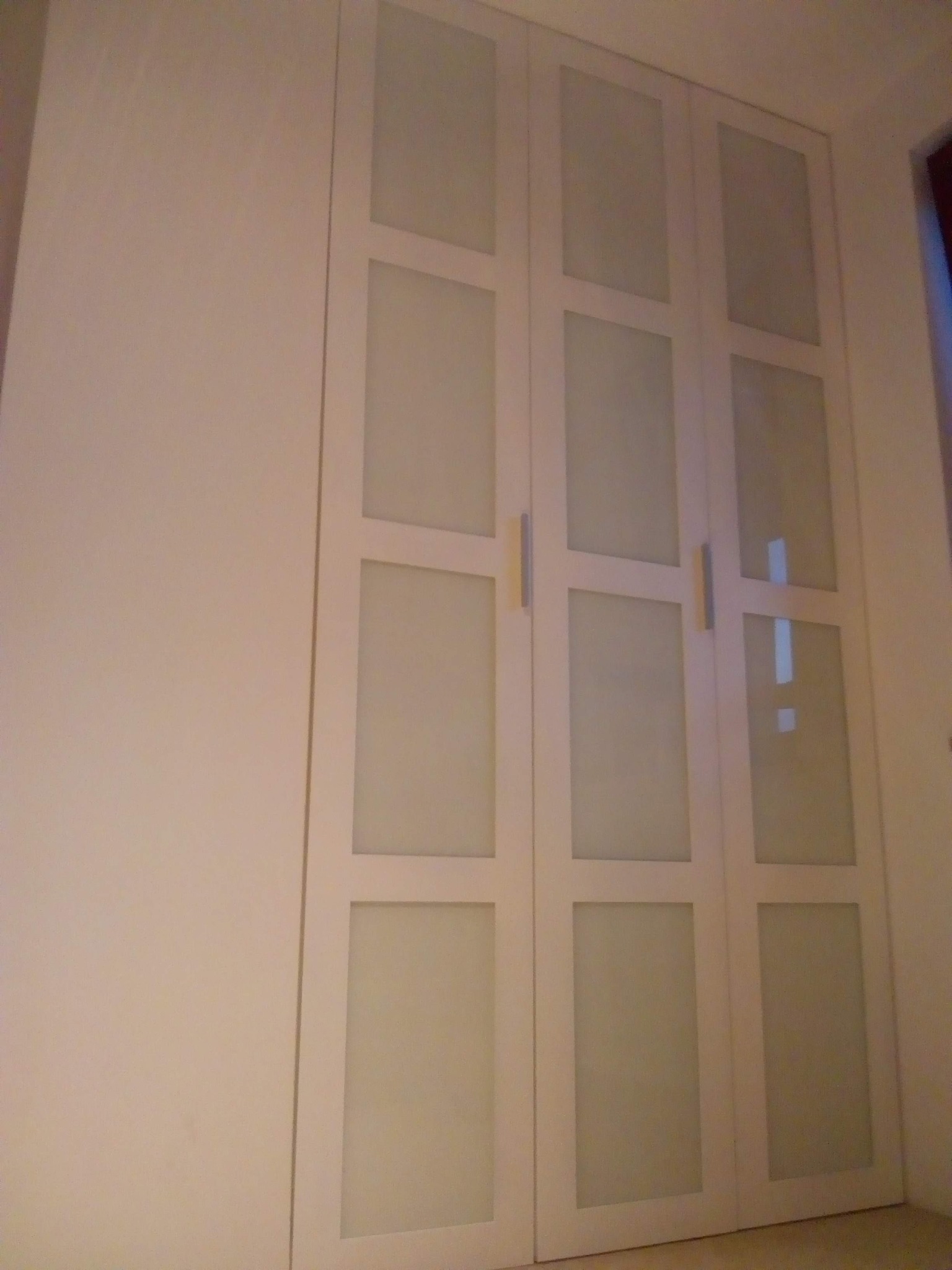 Biała szafa wnękowa z przesuwnymi drzwiami z matowego szkła i podłużnymi uchwytami, umieszczona we wnęce pomieszczenia.