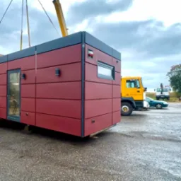 Czerwono-szary dom modułowy transportowany dźwigiem na placu budowy, widoczne ciężarówki i pojazdy w tle. Budynek z oknem i drzwiami w nowoczesnym stylu.