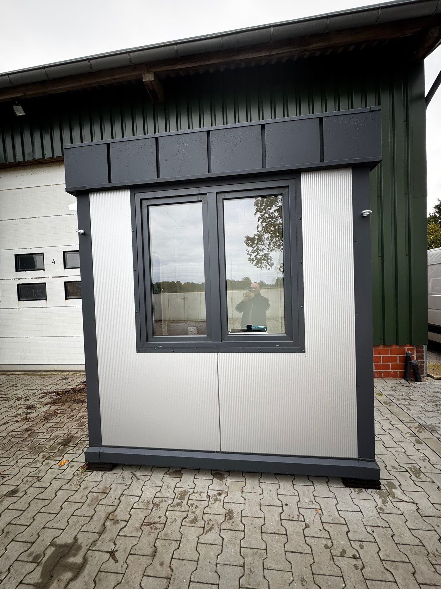 Gotowy, szary pawilon modułowy z oknem na kostce brukowej, z widocznym odbiciem osoby robiącej zdjęcie. W tle budynek z blachy trapezowej i brama garażowa.