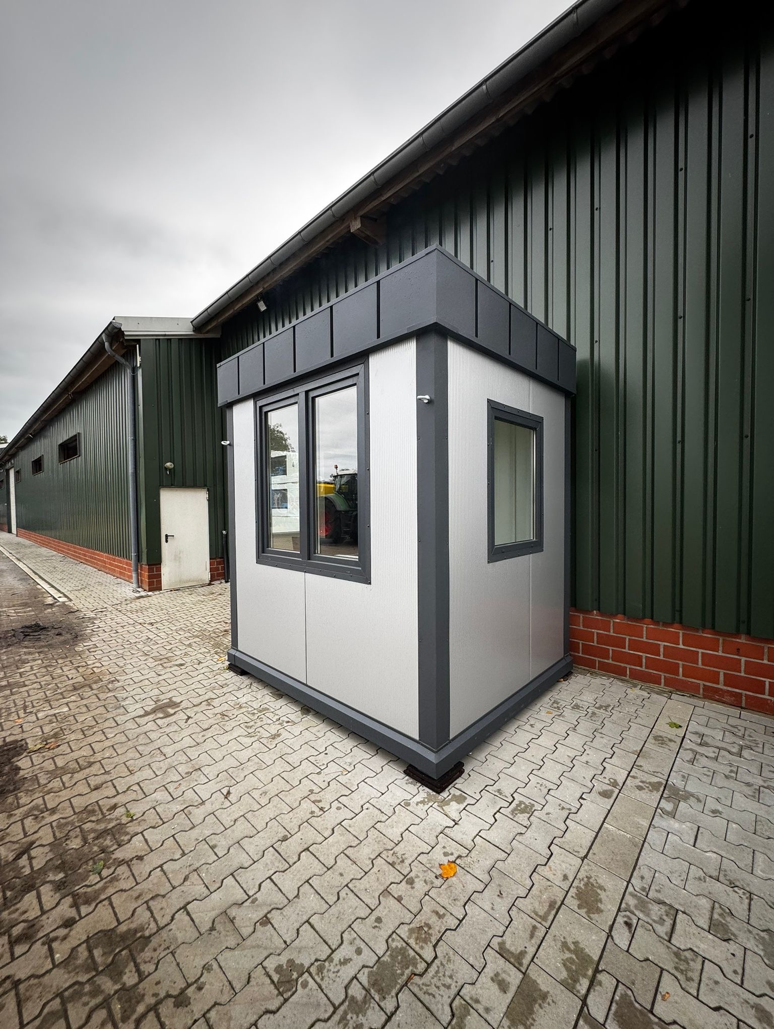 Nowoczesny, srebrny kiosk modułowy z ciemnoszarym wykończeniem, ustawiony na kostce brukowej przed zielonym budynkiem z blachy falistej. W oknach odbija się traktor.