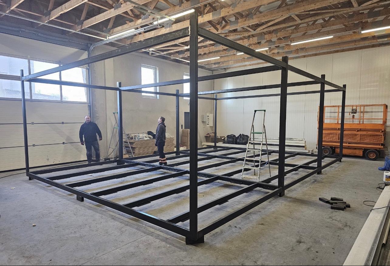 Szkielet stalowej konstrukcji hali w hali produkcyjnej, z widocznymi pracownikami i podnośnikiem. Konstrukcja pomalowana na czarno, zespawana z profili.