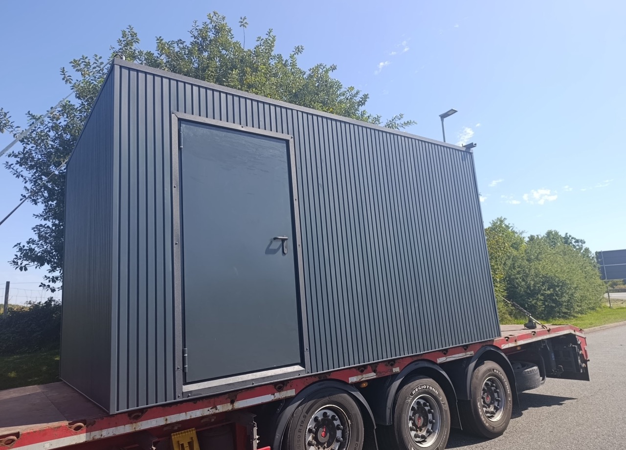 Szary, modułowy budynek z drzwiami, umieszczony na czerwonej przyczepie transportowej. Widoczne drzewa w tle i błękitne niebo. Budynek z blachy trapezowej.