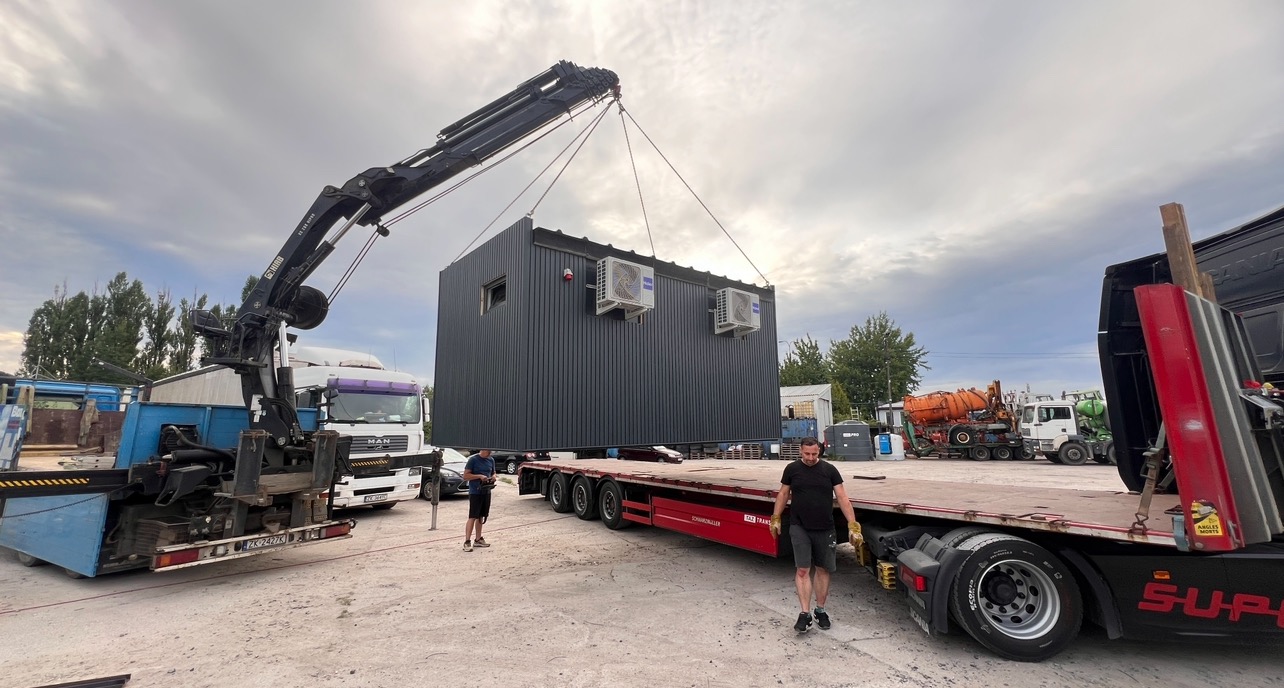 Transport modułowego budynku z klimatyzacją za pomocą dźwigu na naczepie ciężarówki, widoczni pracownicy i inne pojazdy na placu budowy.