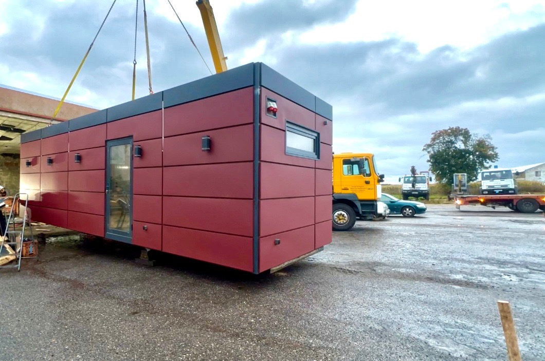 Czerwono-szary dom modułowy transportowany dźwigiem na placu budowy, widoczne ciężarówki i pojazdy w tle. Budynek z oknem i drzwiami w nowoczesnym stylu.