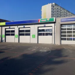 Fasada warsztatu samochodowego z trzema bramami wjazdowymi oznaczonymi numerami 1, 2, 3 i logo Castrol. Obok stacja kontroli pojazdów.