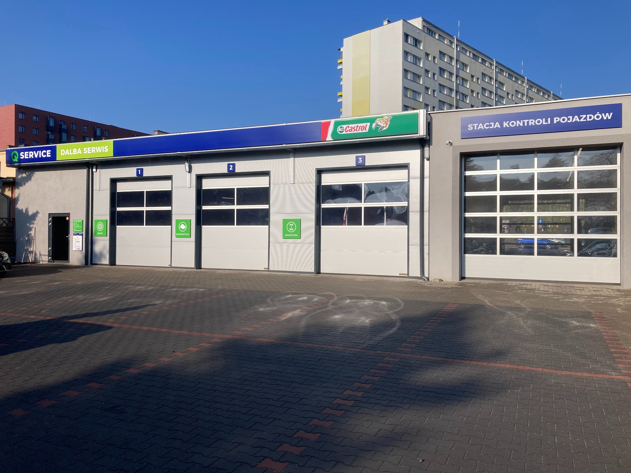 Fasada warsztatu samochodowego z trzema bramami wjazdowymi oznaczonymi numerami 1, 2, 3 i logo Castrol. Obok stacja kontroli pojazdów.