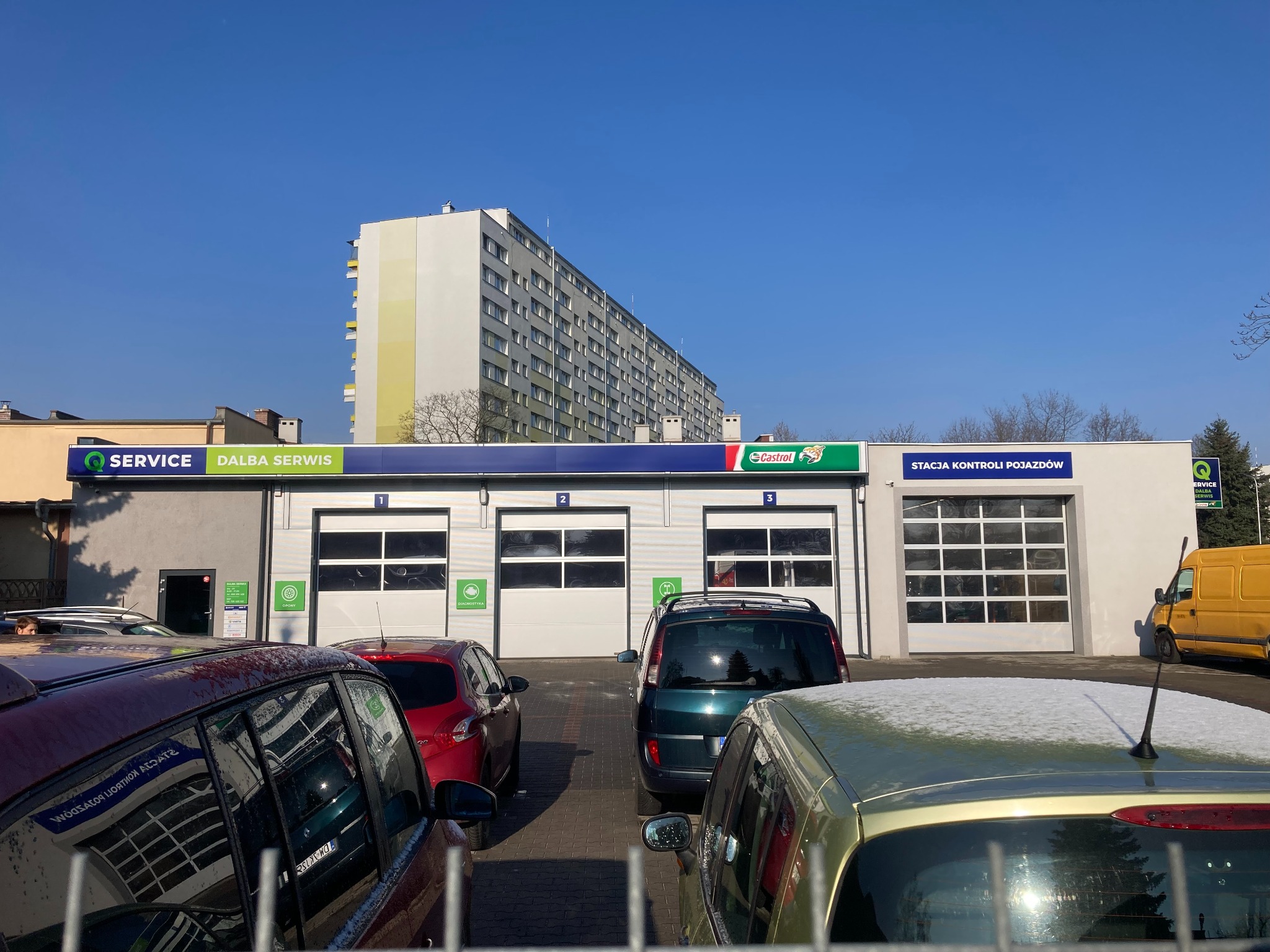 Stacja kontroli pojazdów Dalba Serwis z trzema bramami wjazdowymi i zaparkowanymi samochodami. Na dachu reklama Castrol. W tle wysoki budynek mieszkalny.