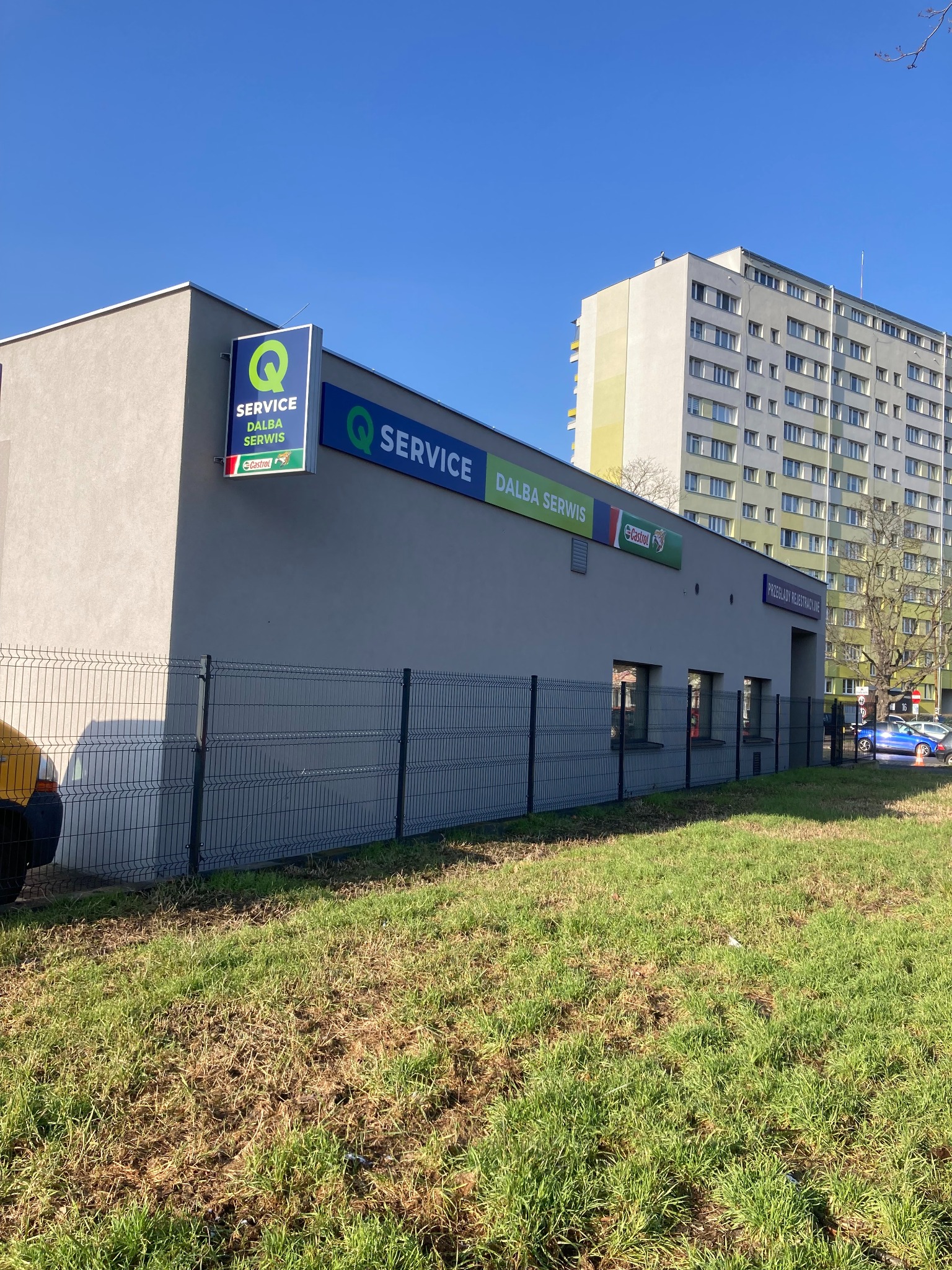 Budynek serwisu samochodowego Dalba Serwis z logo Q Service i Castrol, ogrodzony płotem, na tle bloku mieszkalnego w słoneczny dzień.