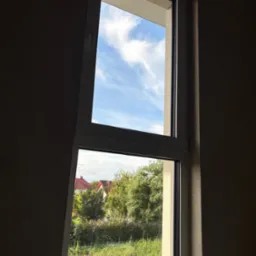 Dwudzielne okno PCV z widokiem na błękitne niebo i zieleń ogrodu.  Perspektywa z wnętrza, ściana w odcieniach beżu.