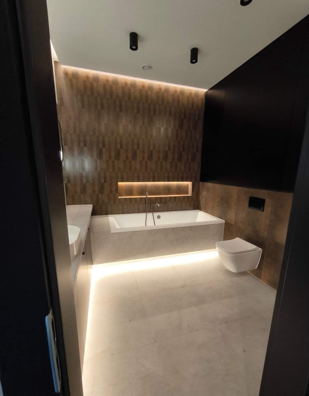 Nowoczesna łazienka z podświetlaną wanną i płytkami imitującymi drewno. Ujęcie zza futryny, widok na minimalistyczną toaletę i umywalkę. Oświetlenie punktowe.