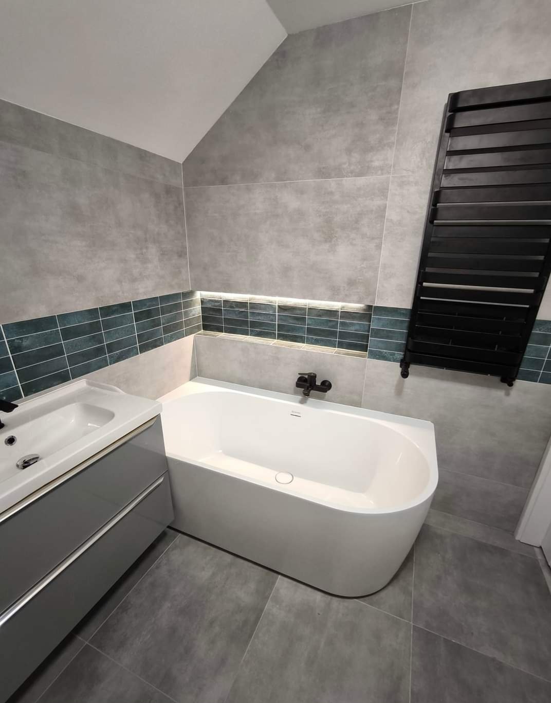Wykończona łazienka z wanną, umywalką i grzejnikiem drabinkowym. Szare płytki na ścianach i podłodze, dekoracyjny pas z podświetleniem LED.