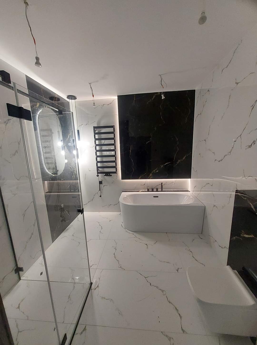 Nowoczesna łazienka z czarno-białymi marmurkowymi płytkami, kabiną prysznicową ze szkła, białą wanną i grzejnikiem drabinkowym. Niedokończone oświetlenie sufitowe.