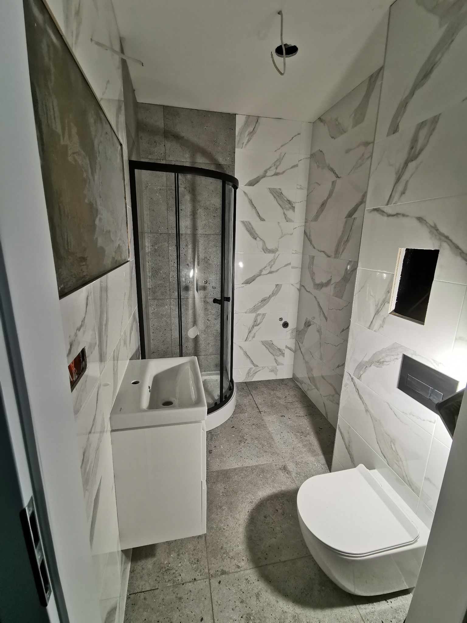 Wykończona łazienka w szaro-białej kolorystyce z kabiną prysznicową z czarnymi ramami, umywalką z szafką i podwieszaną toaletą. Widoczne płytki imitujące marmur.