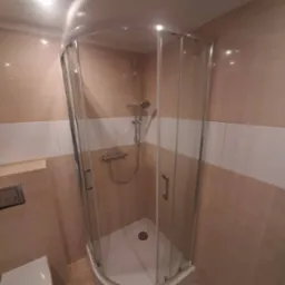 Narożna kabina prysznicowa z chromowanymi profilami i brodzikiem, zamontowana w łazience wykończonej beżowo-białymi płytkami. Widoczna bateria natryskowa i słuchawka.