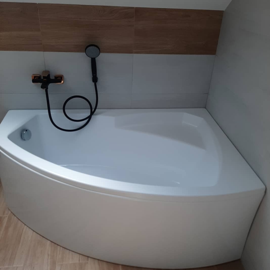 Biała, narożna wanna z czarną słuchawką prysznicową i złotą baterią na tle płytek imitujących drewno. Gotowa łazienka po remoncie.