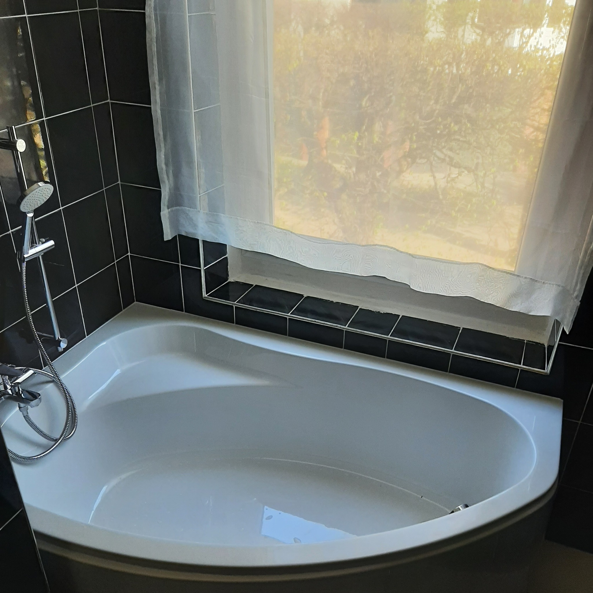 Biała narożna wanna z chromowaną armaturą, obudowana czarnymi płytkami. Okno z firanką w tle. Widok na zieleń za oknem. Ujęcie z góry, oświetlenie naturalne.