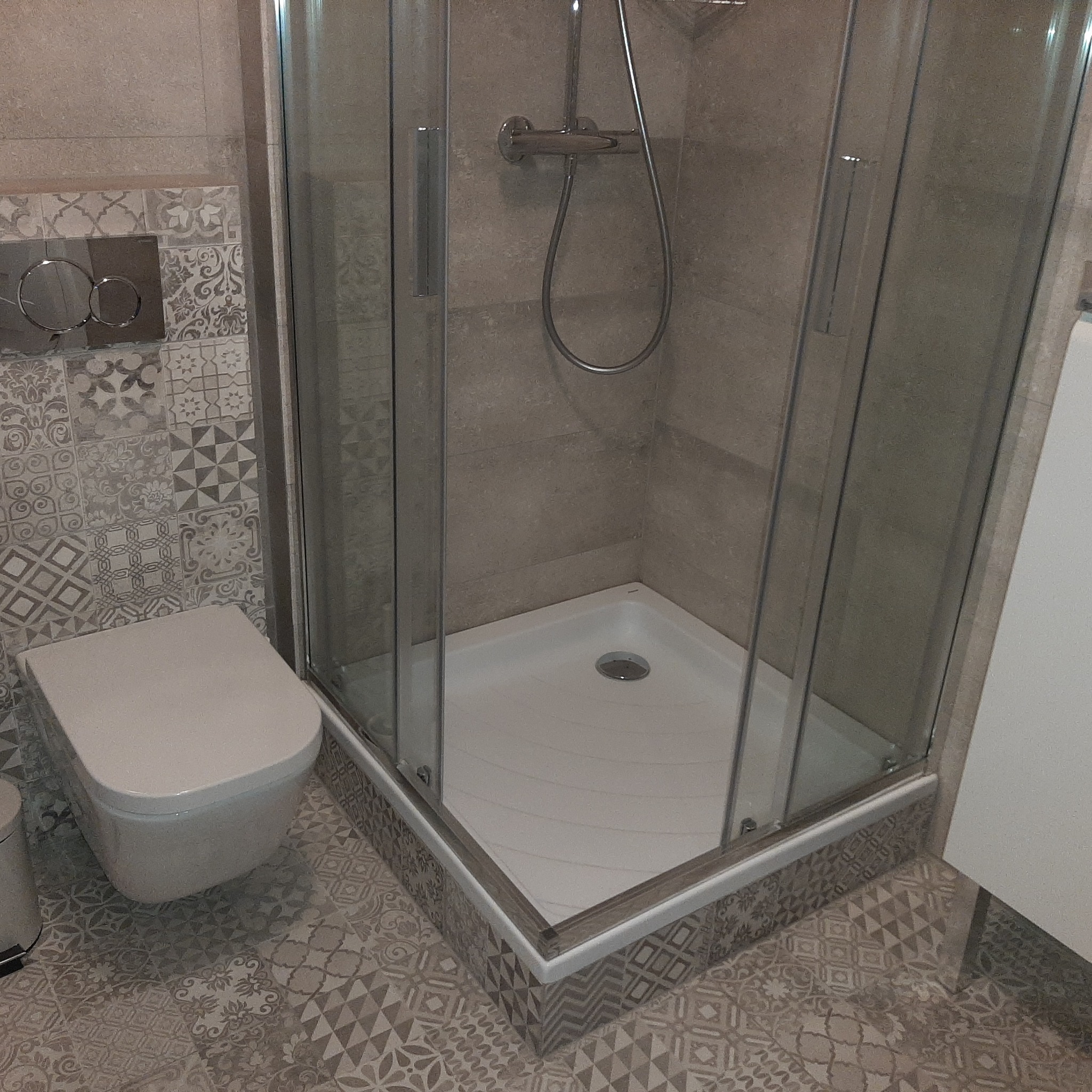 Nowoczesna kabina prysznicowa z deszczownicą w beżowej łazience z mozaiką, obok toaleta wisząca. Widok na detale wykończenia i armaturę.