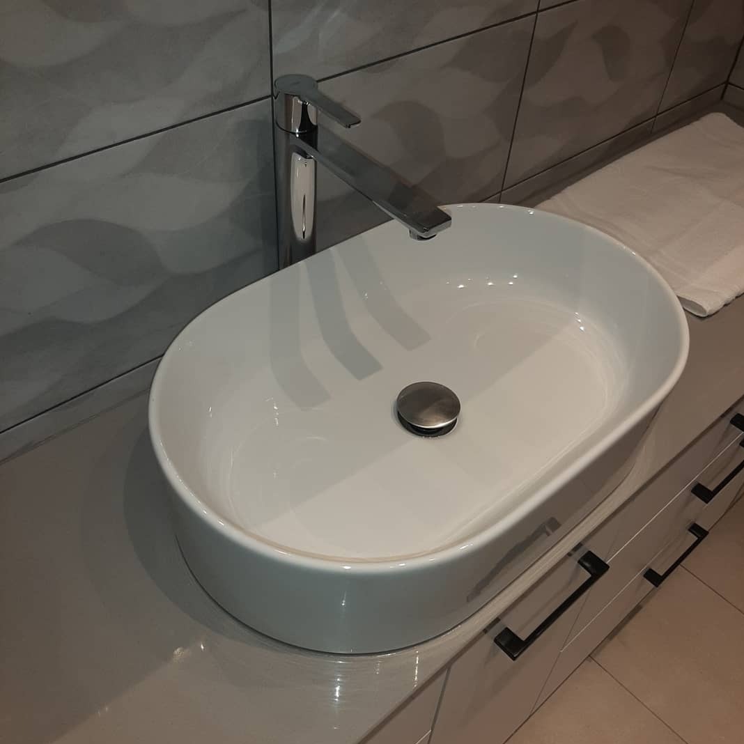 Biała, owalna umywalka nablatowa z chromowaną baterią na szafce z szufladami. W tle szare płytki z delikatnym wzorem i biały ręcznik. Nowoczesna łazienka.