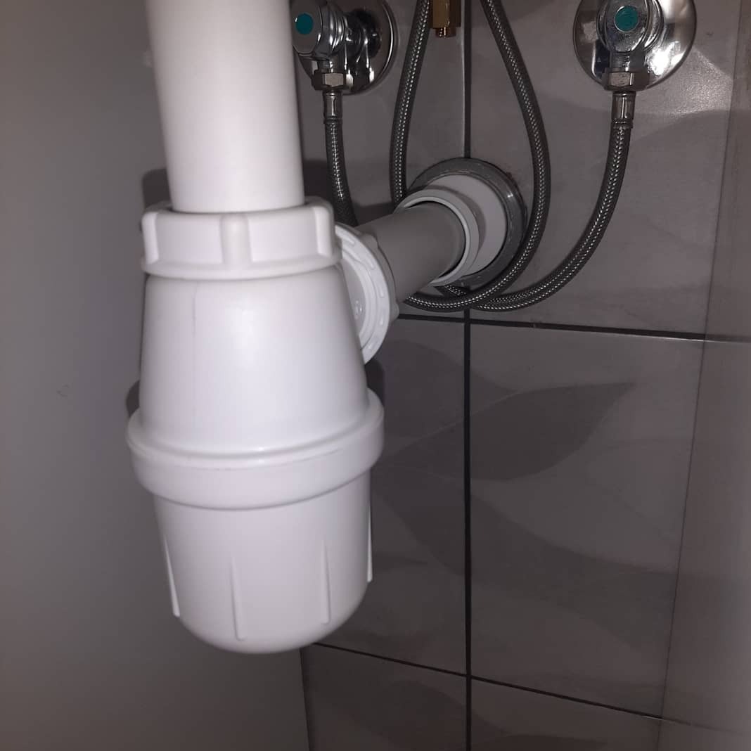 Zamontowany, biały syfon umywalkowy z plastikowymi rurami na tle szarych płytek ceramicznych. Widoczne zawory z wężykami doprowadzającymi wodę.