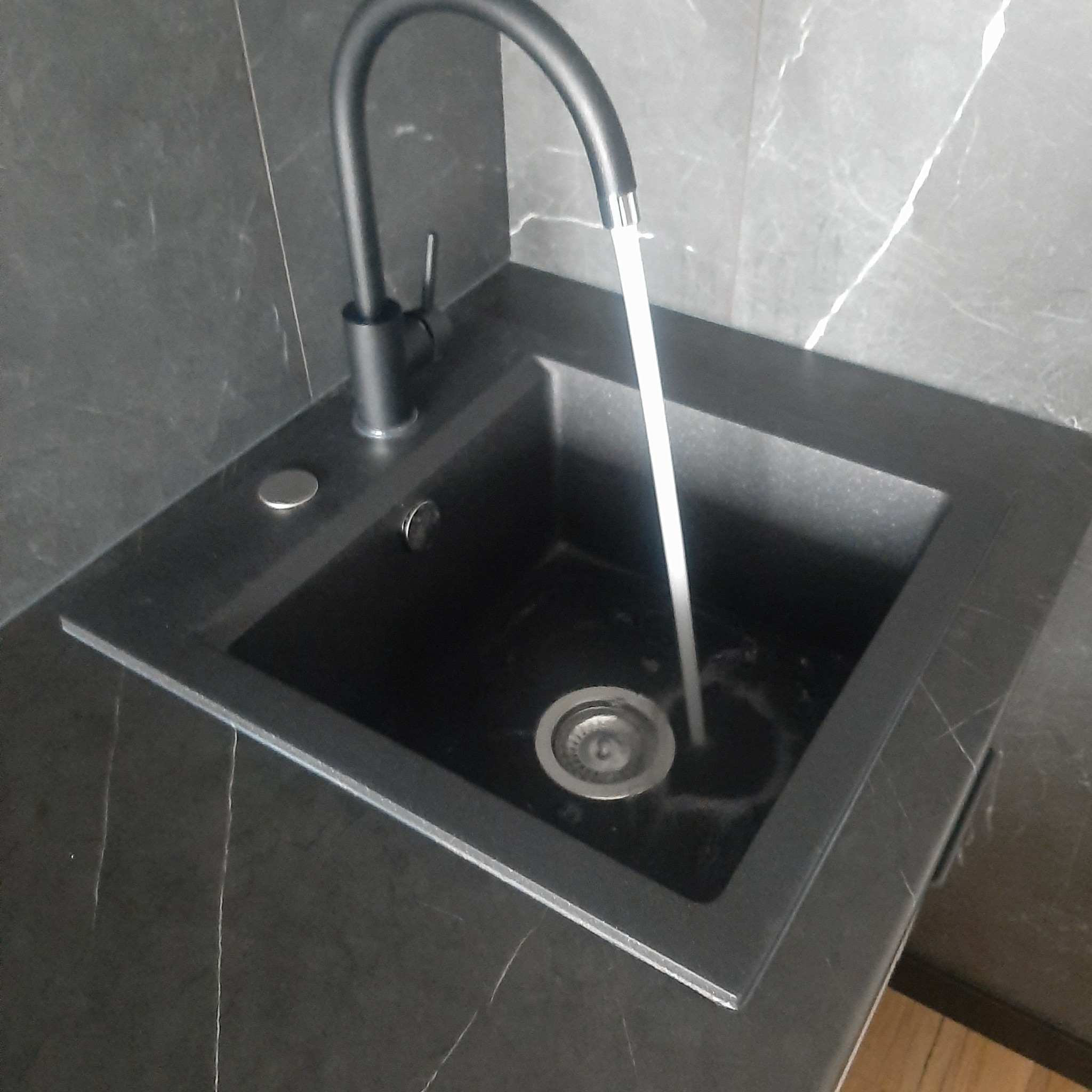 Czarny zlew granitowy z czarną, łukowatą baterią, z której leci woda. Zlew wkomponowany w blat o marmurowej strukturze. Widoczny odpływ i przycisk.