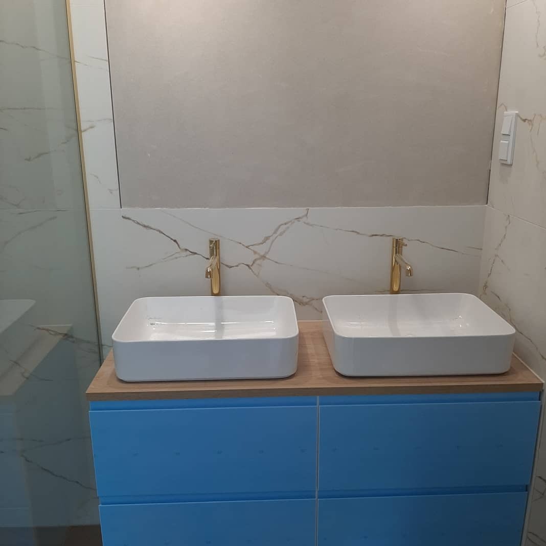 Dwie białe, kwadratowe umywalki z złotymi bateriami na błękitnej szafce w łazience z marmurowymi płytkami. Widoczny fragment kabiny prysznicowej z lewej strony.
