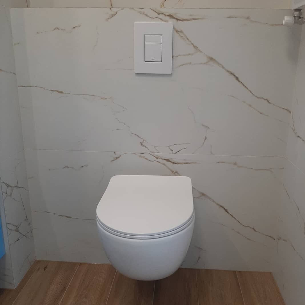 Biała, podwieszana toaleta z przyciskiem spłukiwania na marmurowej ścianie i drewnianą podłogą. Nowoczesna łazienka po remoncie z eleganckim wykończeniem.