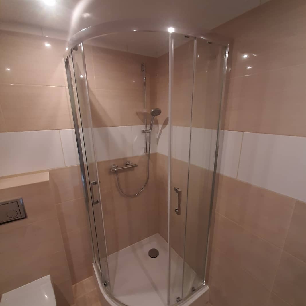 Narożna kabina prysznicowa z chromowanymi profilami i brodzikiem, zamontowana w łazience wykończonej beżowo-białymi płytkami. Widoczna bateria natryskowa i słuchawka.
