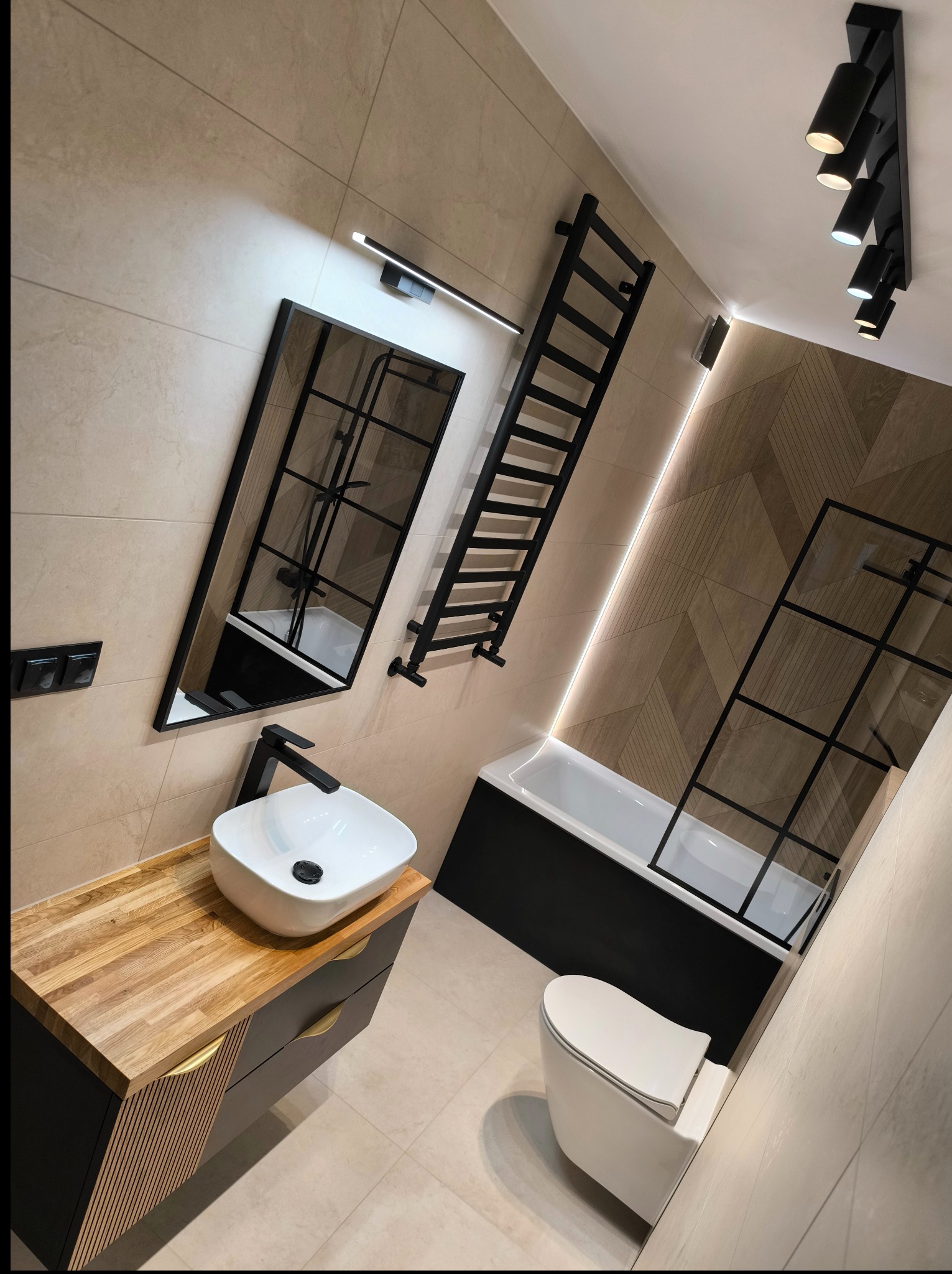 Nowoczesna łazienka z beżowymi płytkami, czarną armaturą i drewnianym blatem pod umywalką. Widok z góry na wannę z parawanem i grzejnik.
