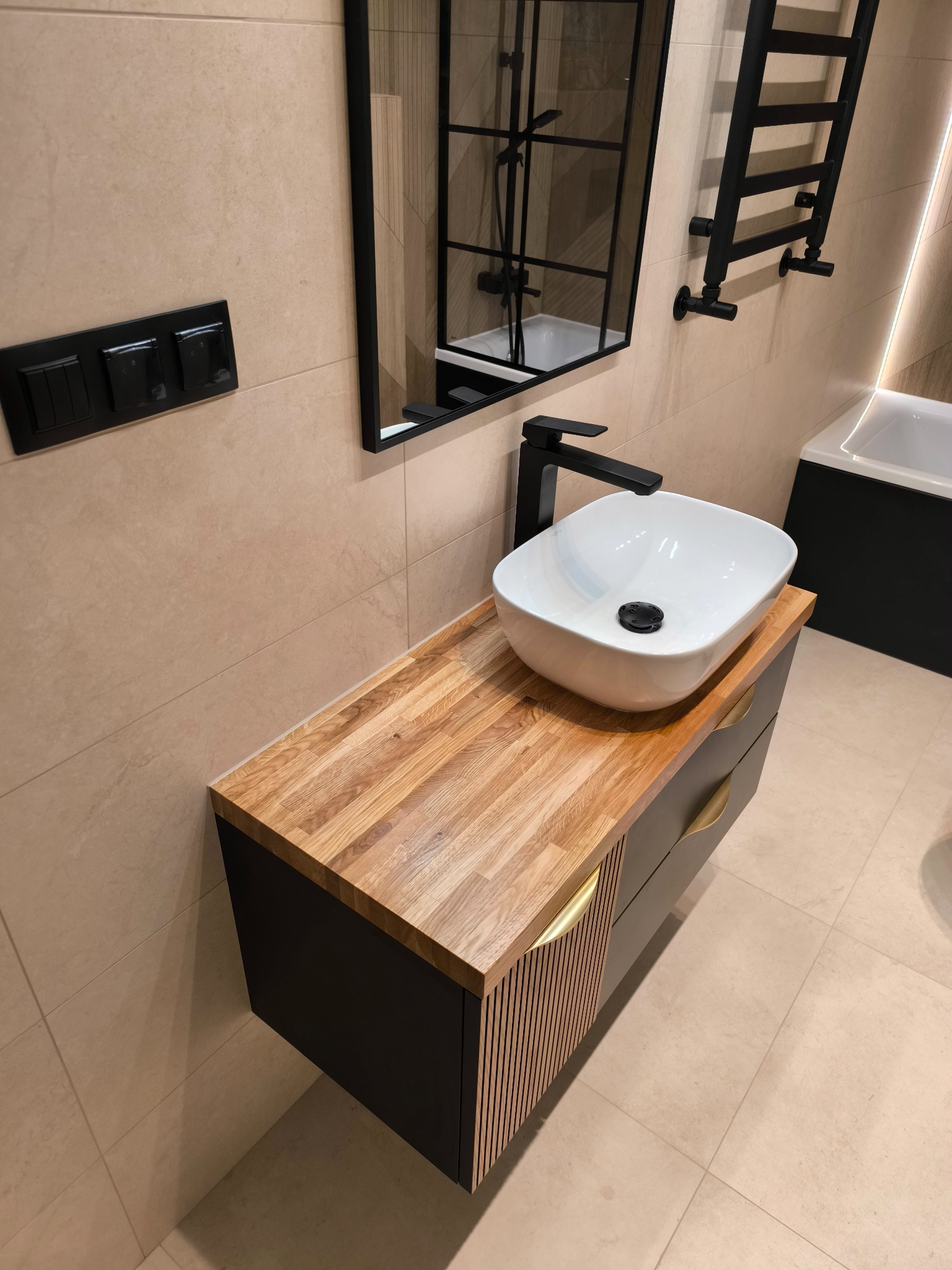 Nowoczesna łazienka: biała umywalka na szafce z drewnianym blatem, czarna armatura, lustro z czarną ramą i grzejnik drabinkowy na beżowych płytkach.
