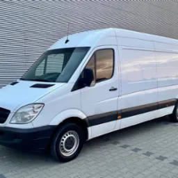 Biały furgon Mercedes-Benz Sprinter z czarnymi akcentami, zaparkowany na kostce brukowej przed budynkiem z szarej blachy falistej.