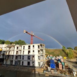 Kamil Dylewski - Budowa bloku z betonu komórkowego w trakcie wznoszenia, na tle tęczy i żurawia budowlanego. Widok z góry, z fragmentem balkonu w kadrze.