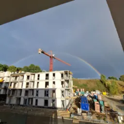 Budowa bloku z betonu komórkowego w trakcie wznoszenia, na tle tęczy i żurawia budowlanego. Widok z góry, z fragmentem balkonu w kadrze.