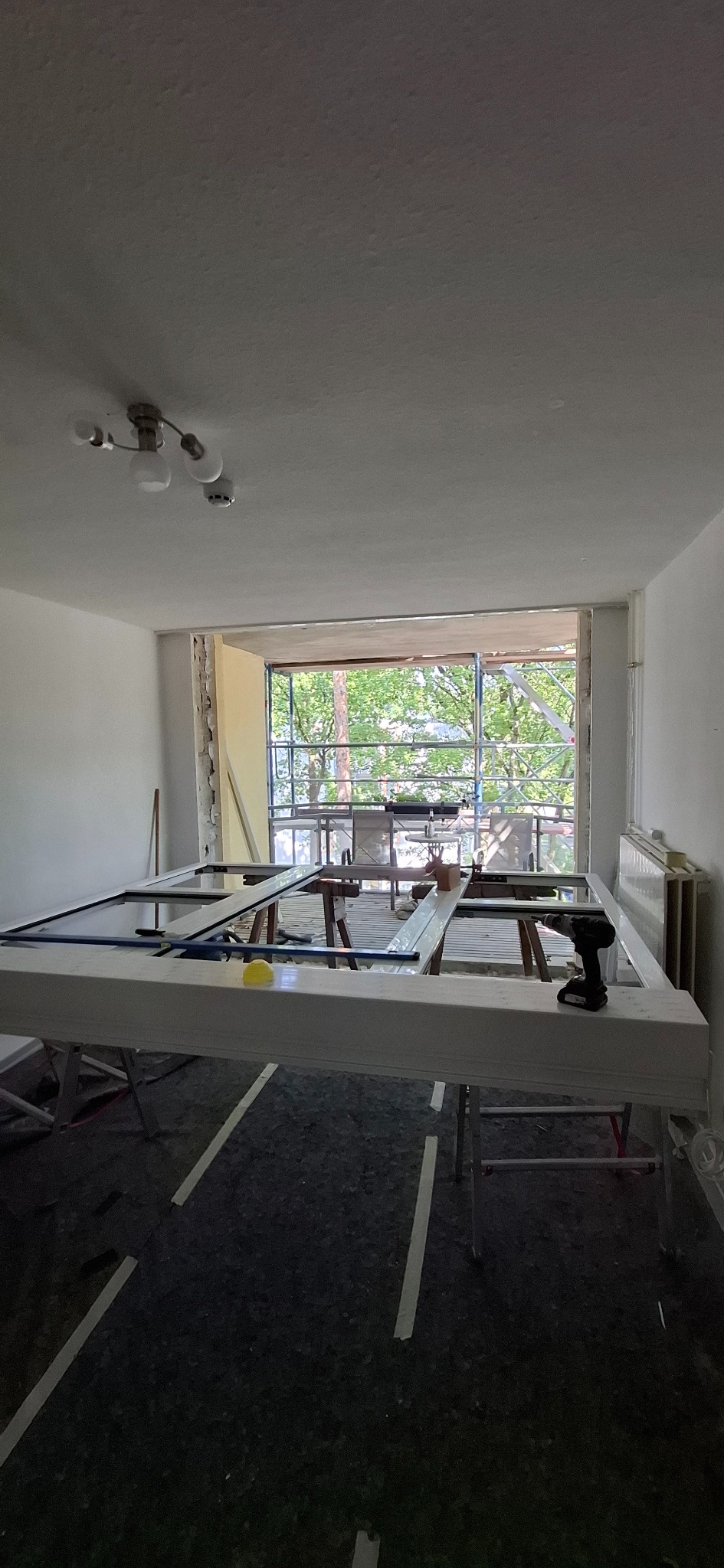 Wnętrze w trakcie remontu z oknem balkonowym, na pierwszym planie rama okienna na kozłach, w tle widok na zieleń, narzędzia i osprzęt budowlany.