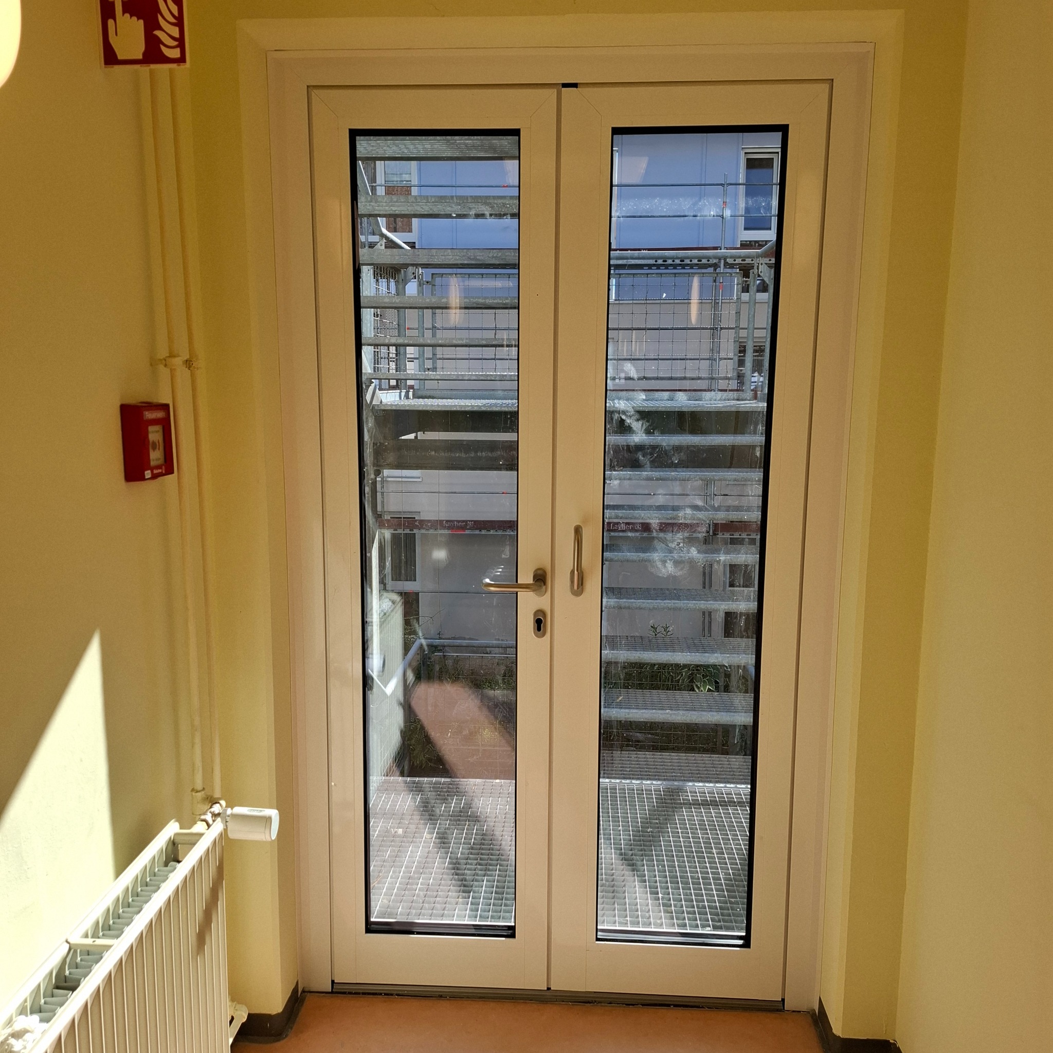Białe, dwuskrzydłowe drzwi balkonowe z widokiem na metalową konstrukcję na zewnątrz, zamontowane w żółtym pomieszczeniu z grzejnikiem i czujnikiem pożarowym.