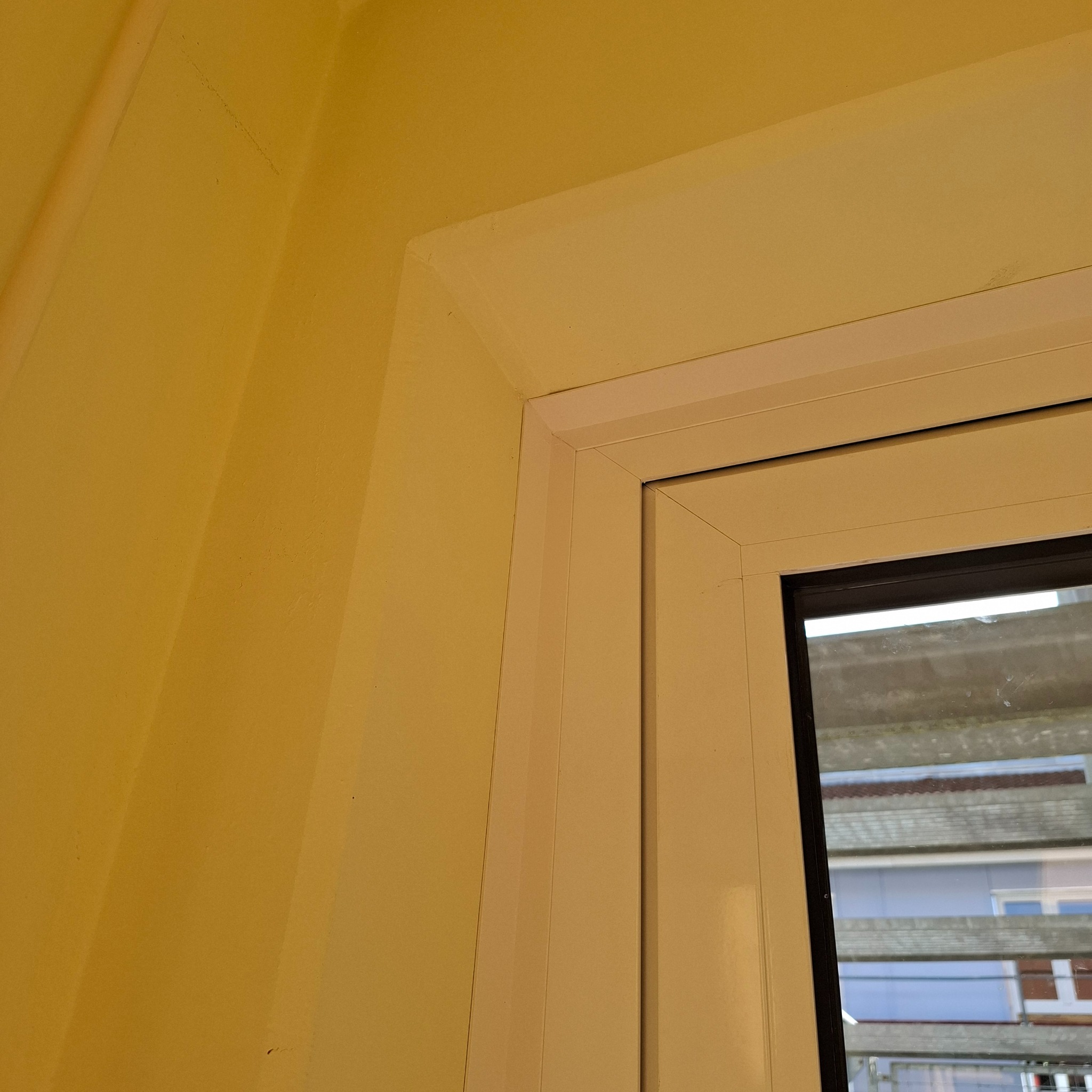 Fragment żółtej ściany z oknem w białej ramie. Widoczne detale wykończenia, kąt ujęcia podkreśla perspektywę wnętrza. Delikatne światło dzienne.