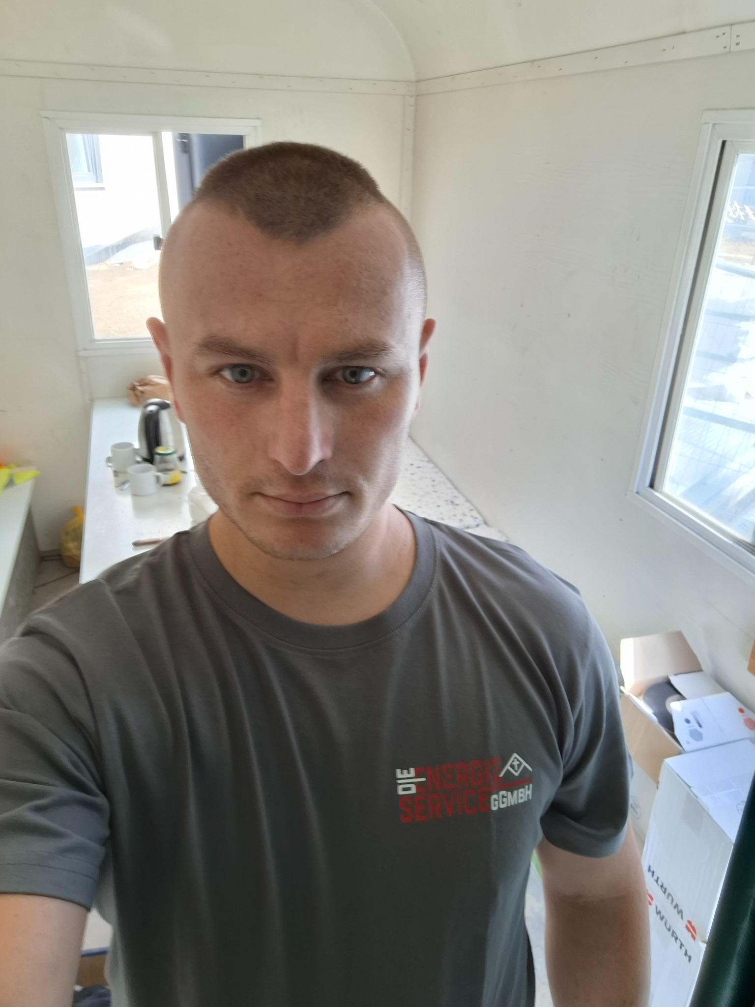 Mężczyzna w szarym t-shircie z logo firmy na tle białego wnętrza z oknami i wyposażeniem biurowym, wykonujący selfie.