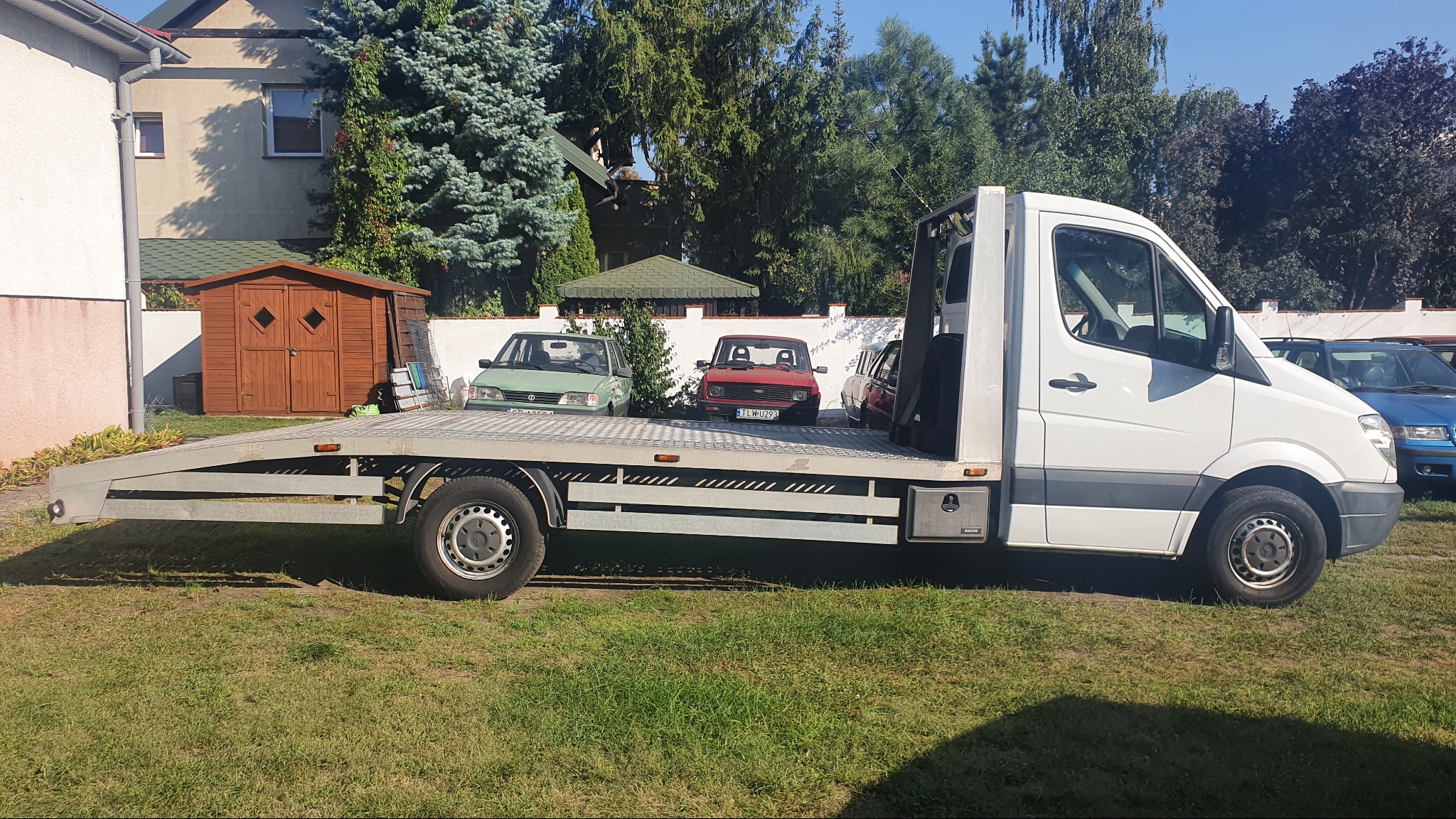 Laweta z trzema samochodami osobowymi na platformie, zaparkowana na trawniku przed domem. Widok z boku, w tle budynek i drzewa.