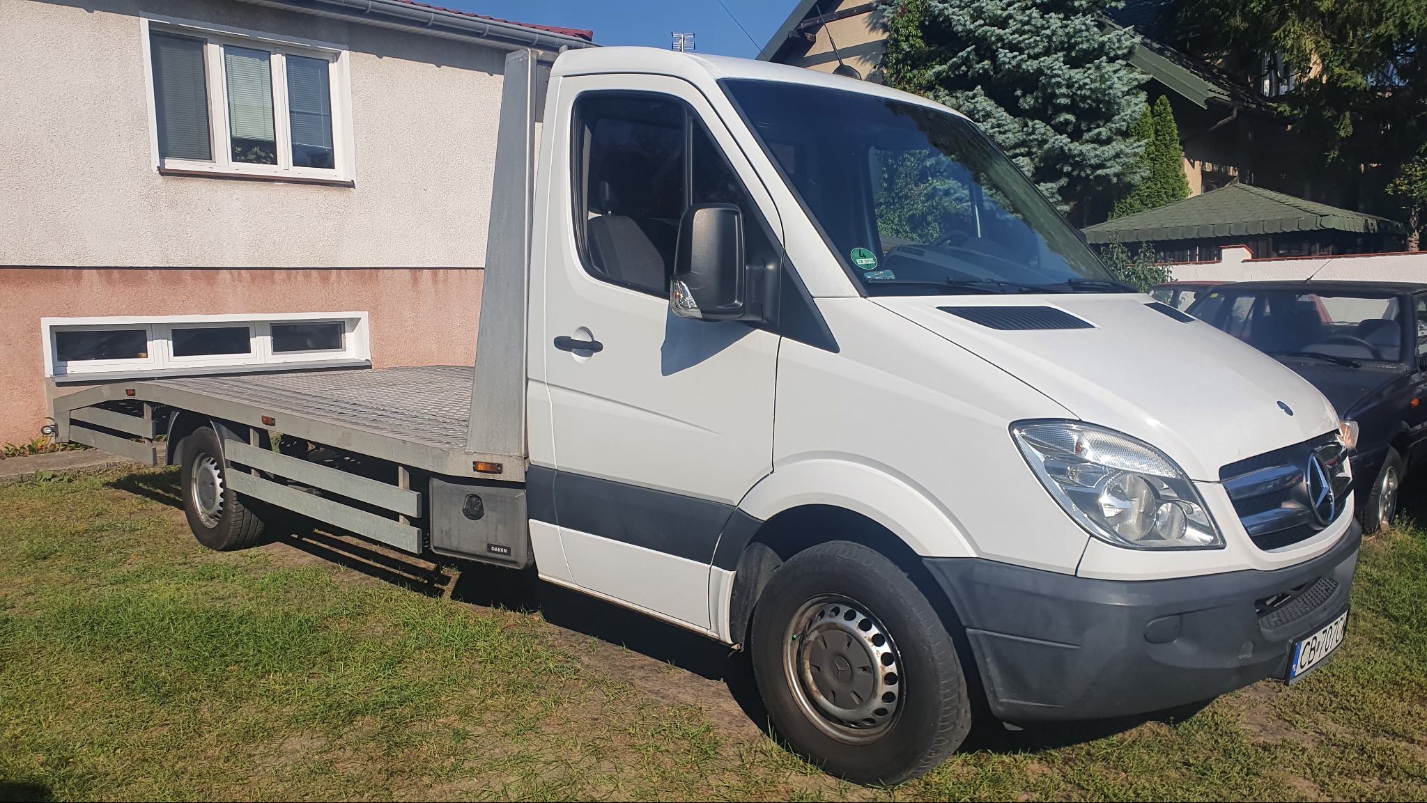 Biała laweta Mercedes Sprinter z kratownicową platformą, zaparkowana na trawie przed budynkiem. Widoczne tablice rejestracyjne i fragment innego pojazdu.