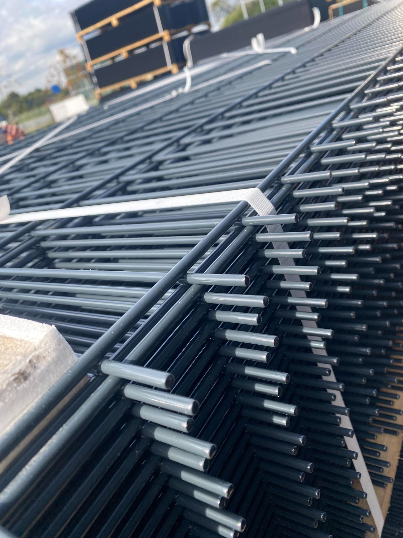 Stos paneli ogrodzeniowych z metalu, zabezpieczonych taśmami, gotowych do transportu. Widoczne elementy mocujące i fragmenty palet.