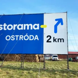 Billboard z logo Castorama i napisem Ostróda 2km na tle błękitnego nieba i zielonego pola. Widoczne bele siana i budynki w oddali. Ujęcie z dołu, podkreślające wielkość reklamy.
