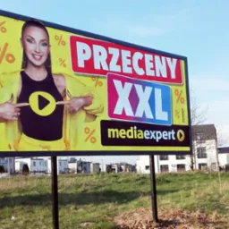 Billboard reklamowy z hasłem 'Przeceny XXL' i wizerunkiem kobiety wskazującej na logo Media Expert, umieszczony na tle osiedla domów jednorodzinnych.