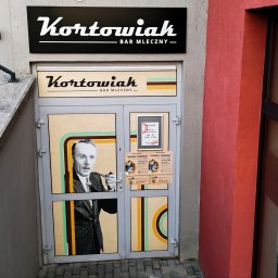 Agencja Reklamowa ActionPrint - Wejście do baru mlecznego Kortowiak z retro grafiką na drzwiach, schody z barierką po lewej, czerwona ściana po prawej, szyld nad wejściem.