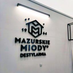 Agencja Reklamowa ActionPrint - Elegancki, czarny logotyp 'Mazurskie Miody Destylarnia' z datą 1964 na białej ścianie, podświetlony listwą LED w Ostródzie. Minimalistyczny design.