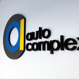 Agencja Reklamowa ActionPrint - Eleganckie logo firmy 'Auto Complex' na białej ścianie. Logo z elementami czerni, błękitu i żółci, obok napis wykonany czarnymi, przestrzennymi literami.