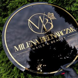 Agencja Reklamowa ActionPrint - Elegancki, okrągły szyld z czarnego akrylu ze złotymi literami 'Milena Bednarczyk Nails & Beauty' i logo z motylem, trzymany na tle zielonych gałązek.