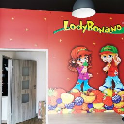 Agencja Reklamowa ActionPrint - Duży, czerwony baner reklamowy z logo 'Lody Bonano' i postaciami dzieci trzymających lody, umieszczony na ścianie, widoczny fragment maszyny do lodów i dwoje drzwi.