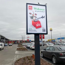 Agencja Reklamowa ActionPrint - Billboard reklamowy sklepu BIG w Ostródzie. Na plakacie kobieta wpadająca do czerwonego pudełka z napisem BIG, hasło: Wskocz po prezenty!, data i kod QR.