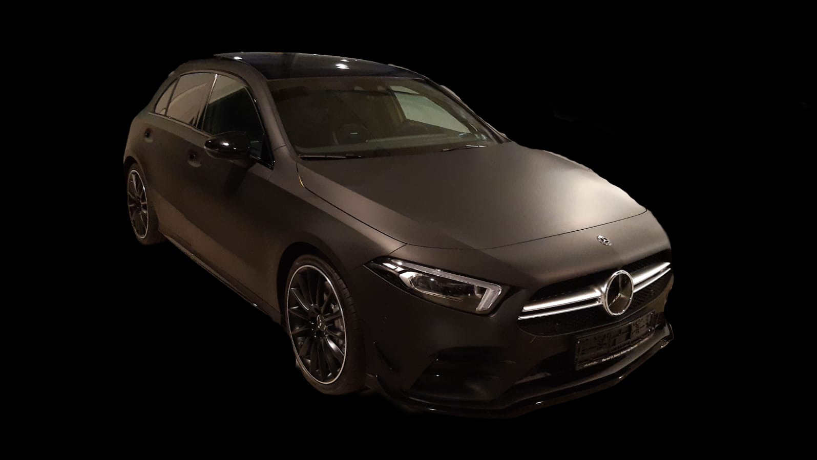 Czarny, matowy Mercedes klasy A na czarnym tle. Widok z przodu, lekko z góry. Reflektory i logo marki są wyraźnie widoczne.